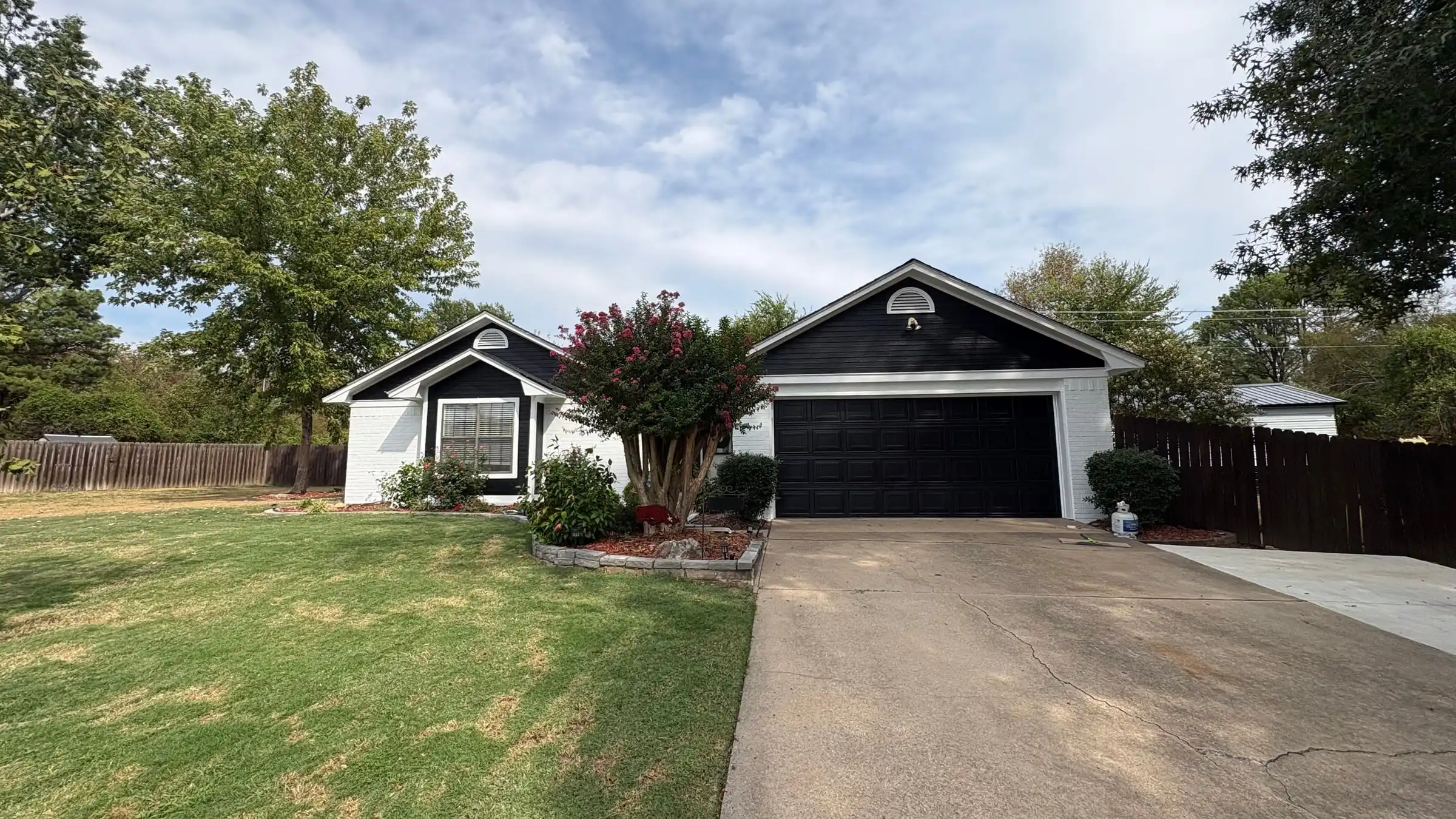 The Fort Smith house from the street after painting: black siding and a black garage door, white brick, a crepe myrtle in bloom beside the drive