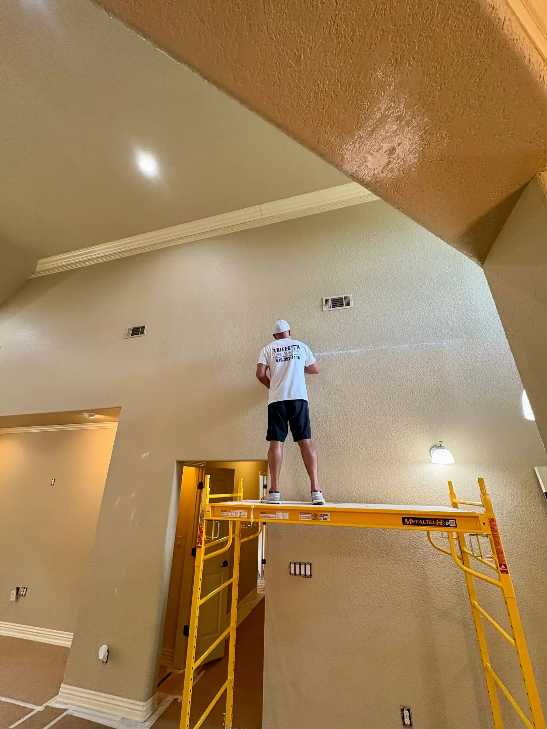 A painter in a Trifecta Pro Painting shirt on a scaffold, working on a tall interior wall