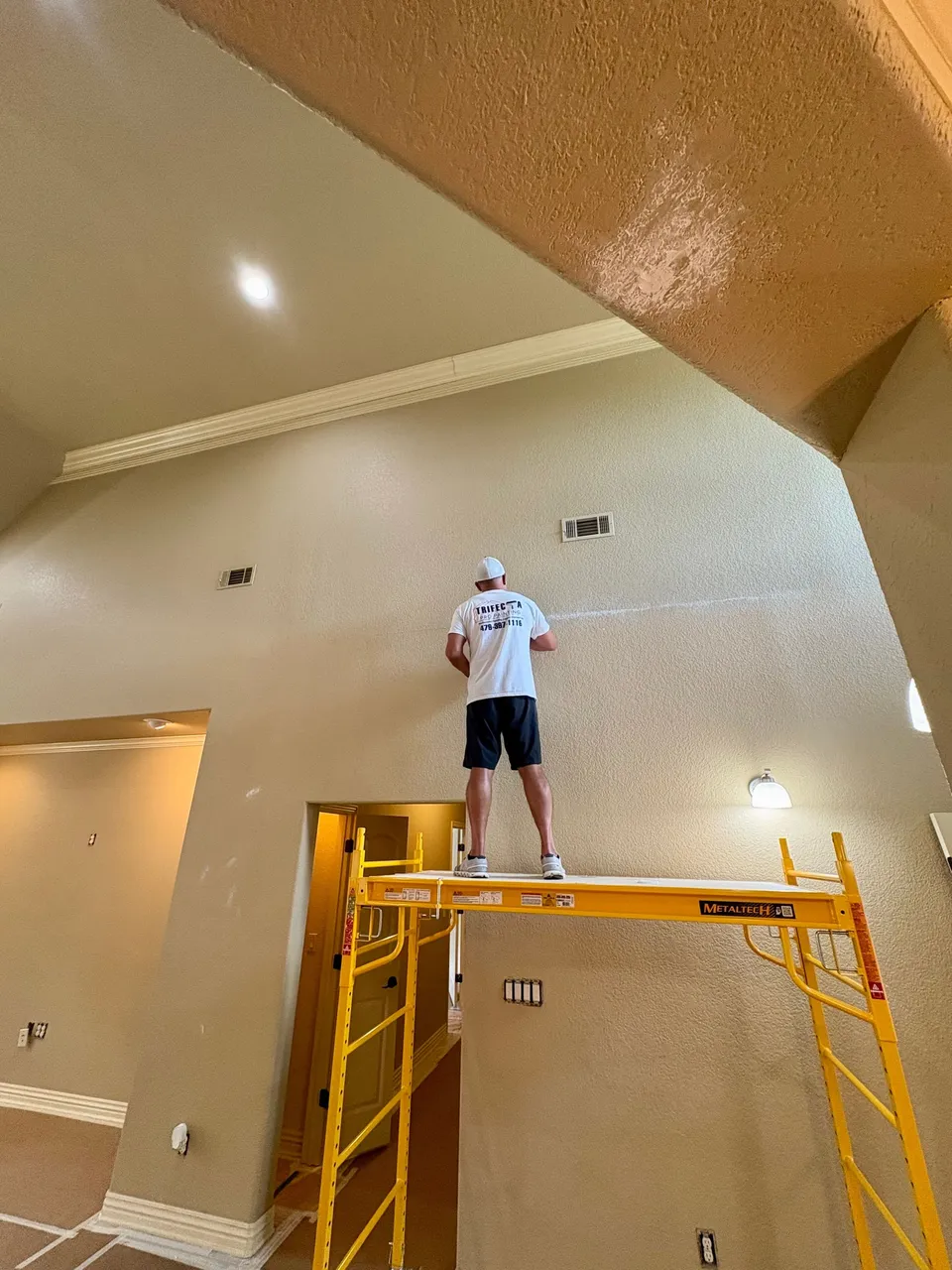 A painter in a Trifecta Pro Painting shirt on a scaffold, working on a tall interior wall