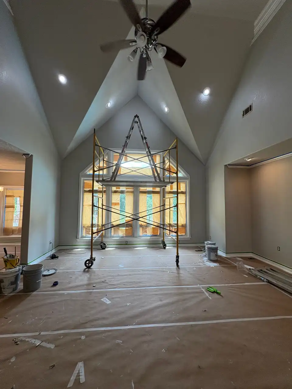 A vaulted great room during painting: a rolling scaffold under the peak, the floor covered in paper and the tall windows masked
