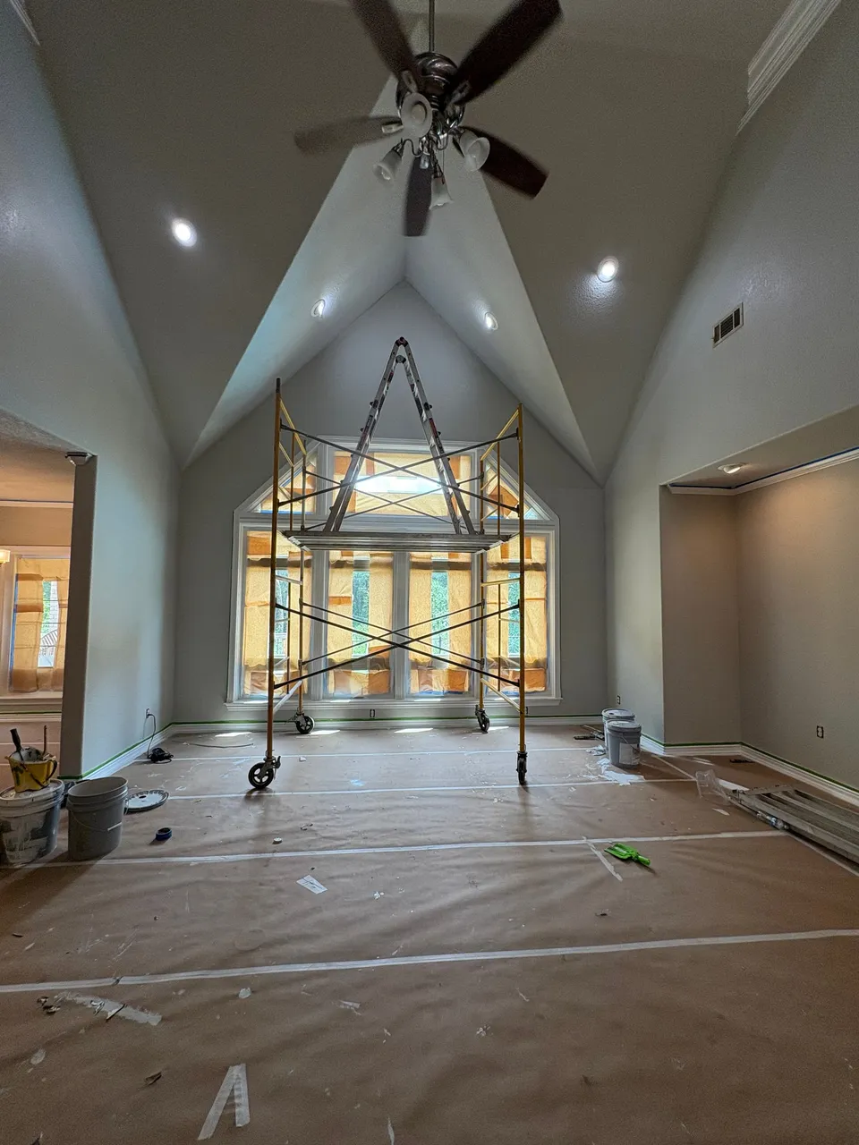 A vaulted great room during painting: a rolling scaffold under the peak, the floor covered in paper and the tall windows masked