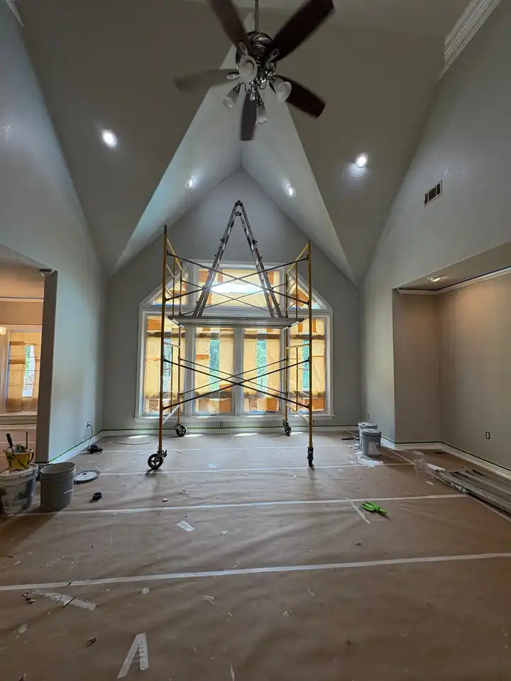 A vaulted great room during painting: a rolling scaffold under the peak, the floor covered in paper and the tall windows masked